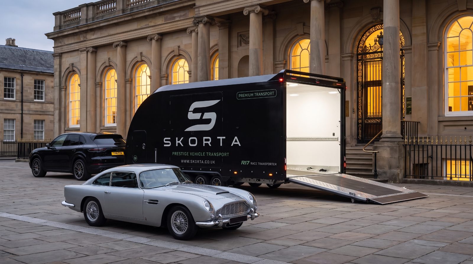 A silver classic grand tourer beside the open SKORTA trailer outside an auction house